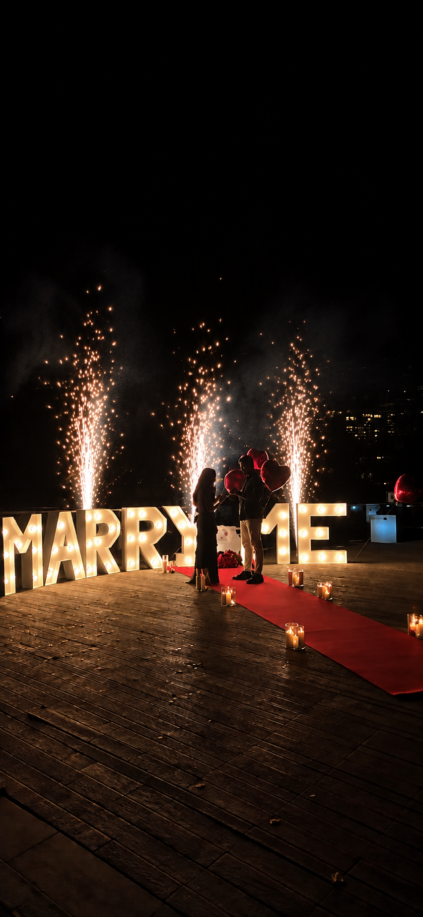 Paar auf rotem Teppich vor leuchtenden Marry-Me-Buchstaben und Fontänen bei Nacht